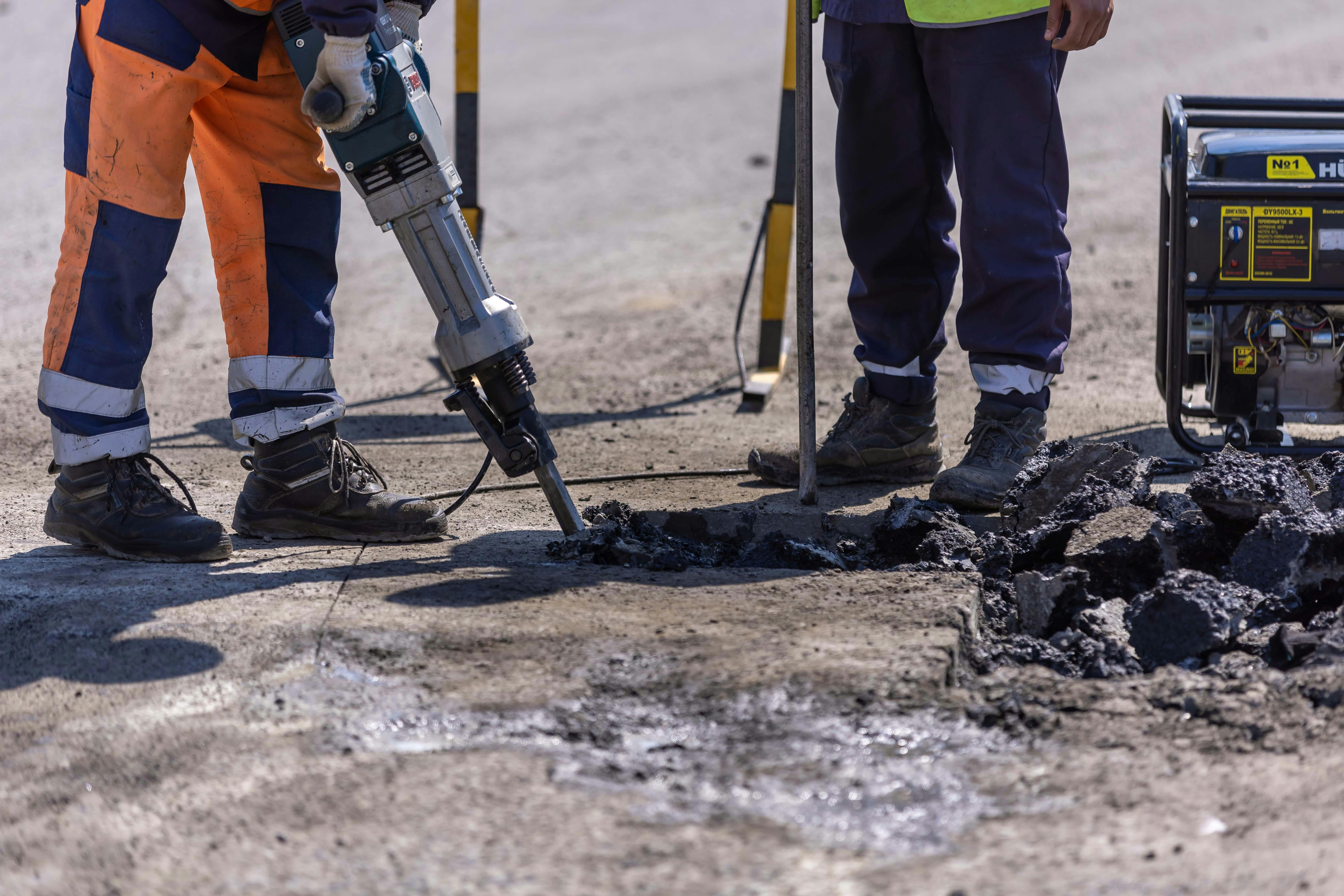 Man being shown using a breaker drill on a concrete ground outdoors with another man standing beside him
